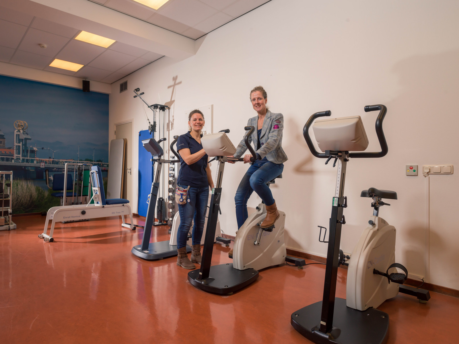 Een persoon fietst op een hometrainer, medewerker staat er naast. Beiden kijken naar de camera.