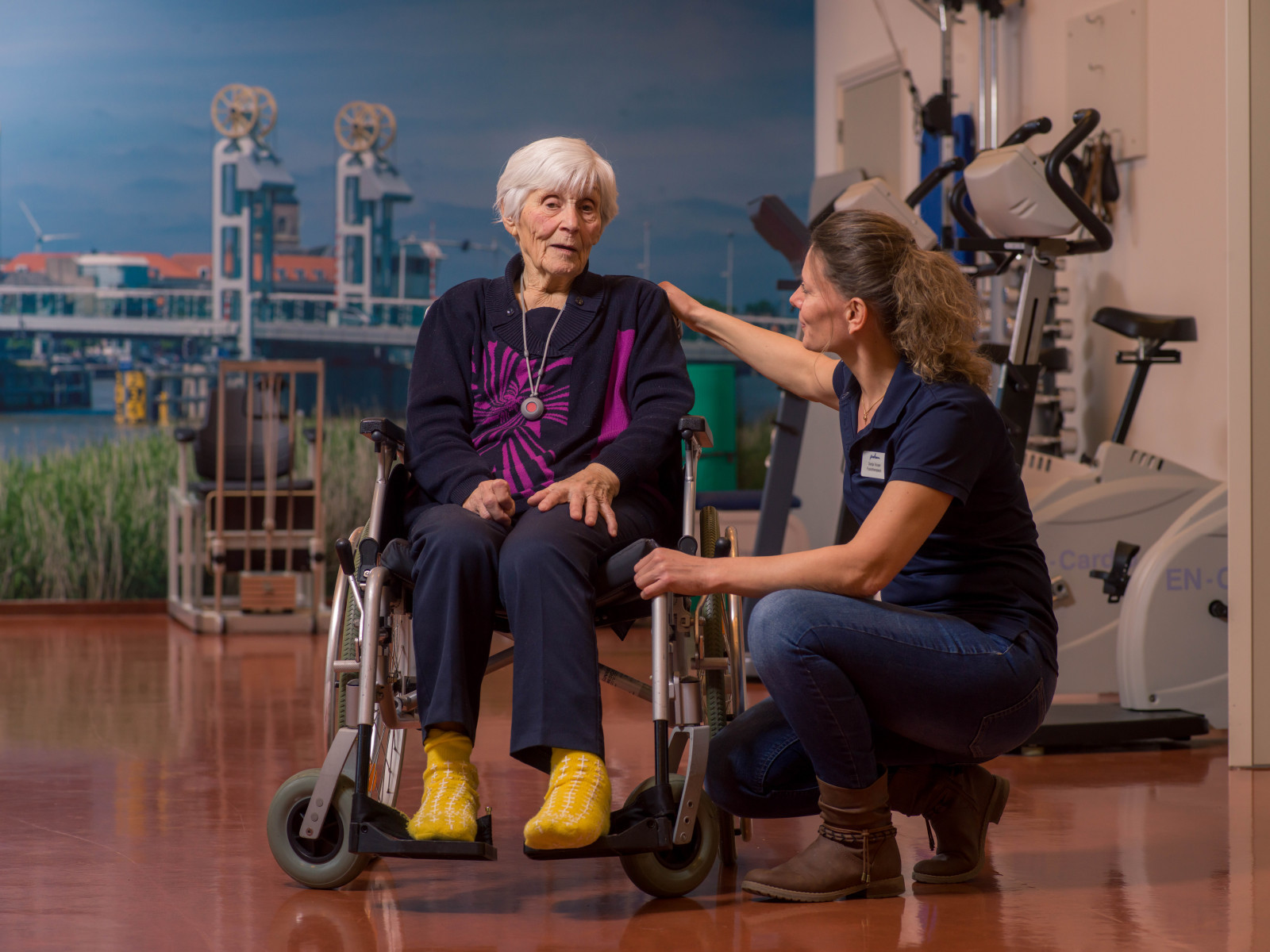 Medewerker van IJsselheem bukt naast de cliënt in rolstoel in een revalidatiezaal.