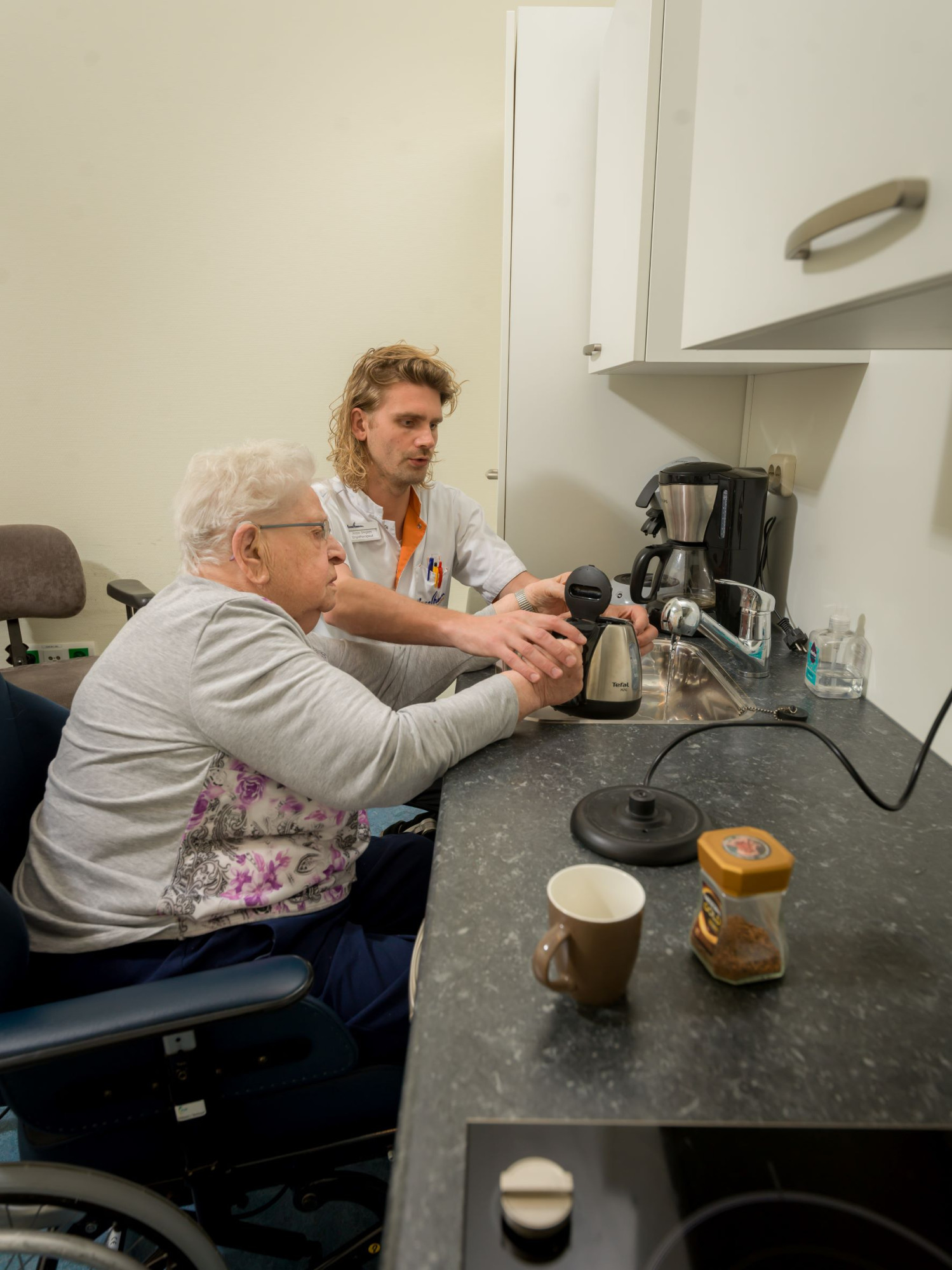Medewerker helpt cliënt met een theekoker vullen onder de kraan